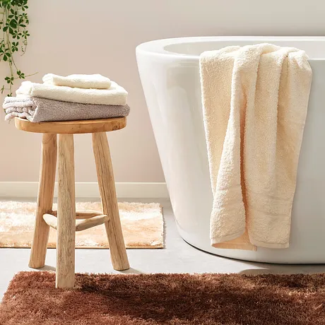 Serviettes de toilette douces en couleur crème et en tons de beige posées sur la baignoire et sur un tabouret dans une salle de bain claire.