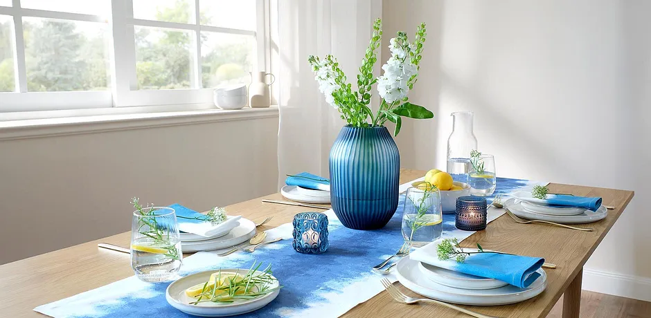 Un chemin de table bleu décore la table de salle à manger, en contraste avec des assiettes blanches et des accessoires colorés.