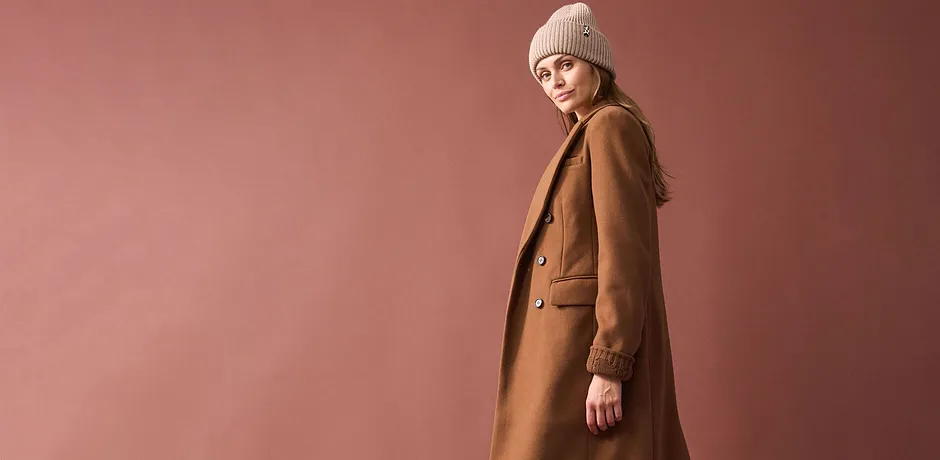 Manteau long marron camel, bonnet en maille beige et une tenue simple en dessous.