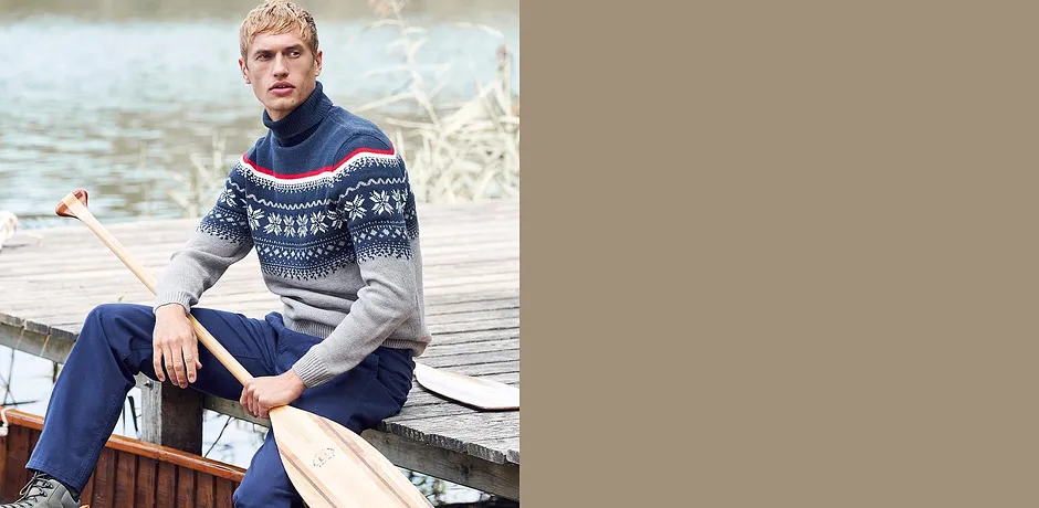 Un homme vêtu d'un pull à motif norvégien et d'un jean, assis sur une berge.