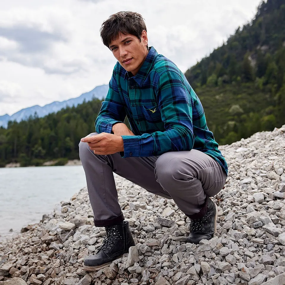 Un randonneur vêtu d'une chemise à carreaux et d'un pantalon gris assis au bord d'une rivière dans un paysage de montagne. 
