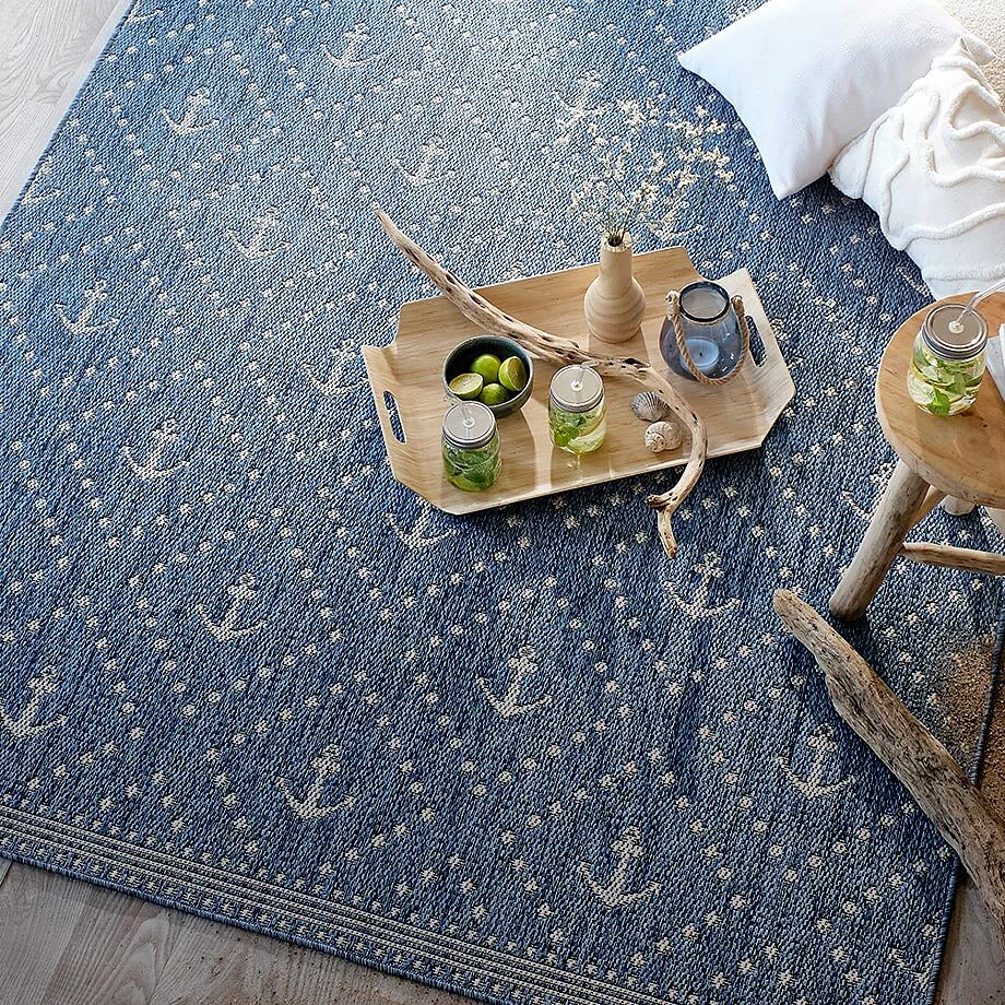 Tapis bleu &agrave; motif ancre marine, coussins blancs et boissons dispos&eacute;es sur un plateau en bois. 