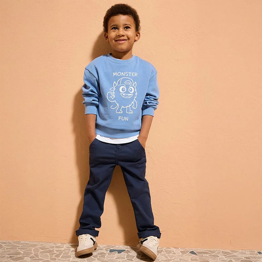 Hellblauer Pullover mit wei&szlig;em Aufdruck, dunkle Jeans und beige Schuhe bilden ein l&auml;ssiges Outfit.