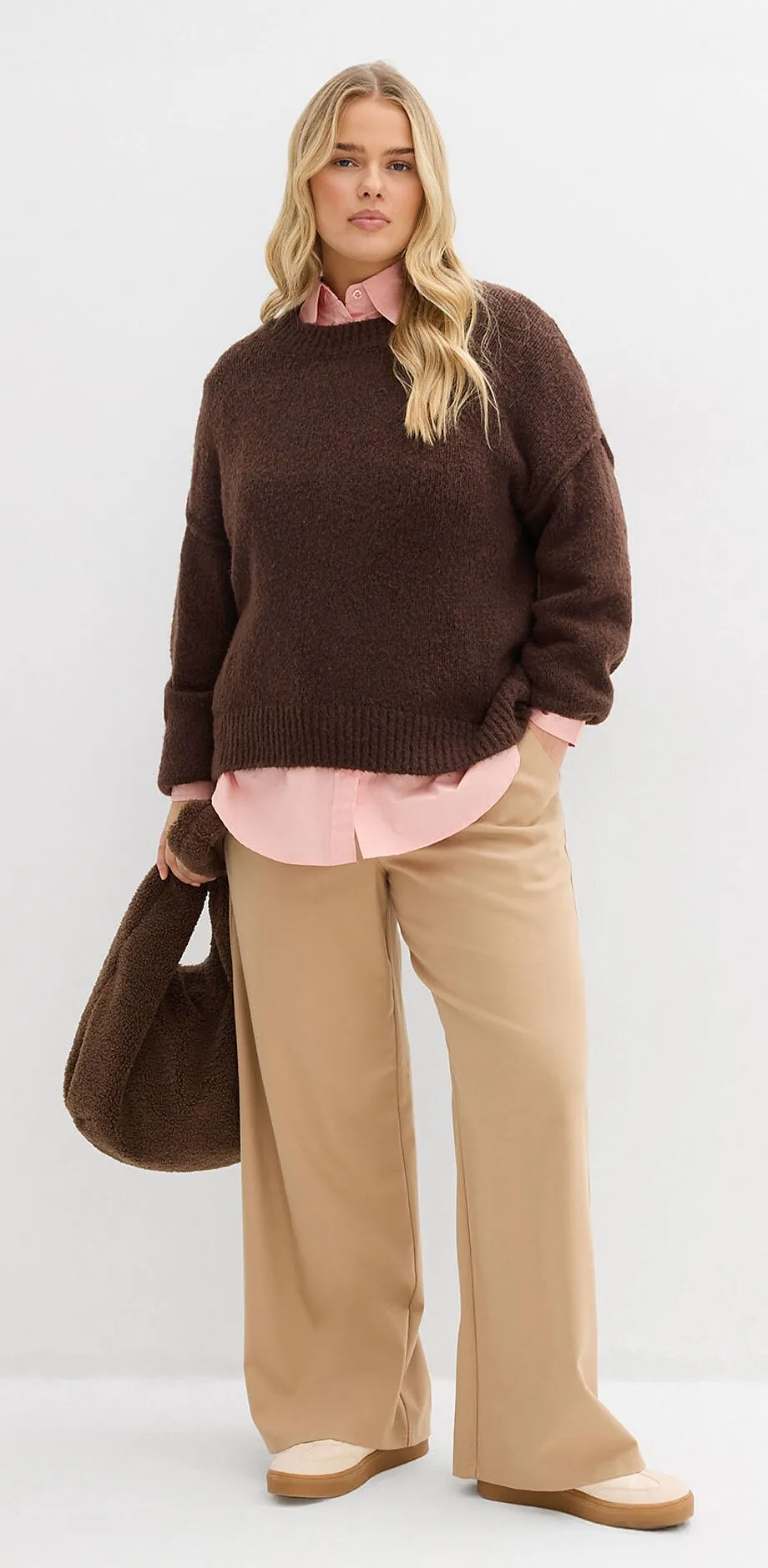 Pull marron par dessus une chemise rose, pantalon beige et sac &agrave; main marron.