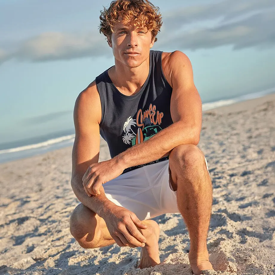 Mann tr&auml;gt ein blaues Tanktop mit Motiv und wei&szlig;e Shorts am Strand.