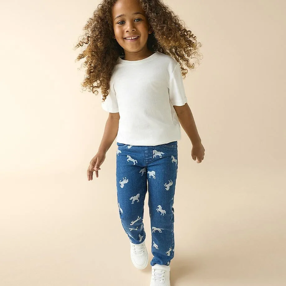 Petite fille v&ecirc;tue d'un T-shirt blanc, un jean bleu avec motifs chevaux et sneakers blanches.