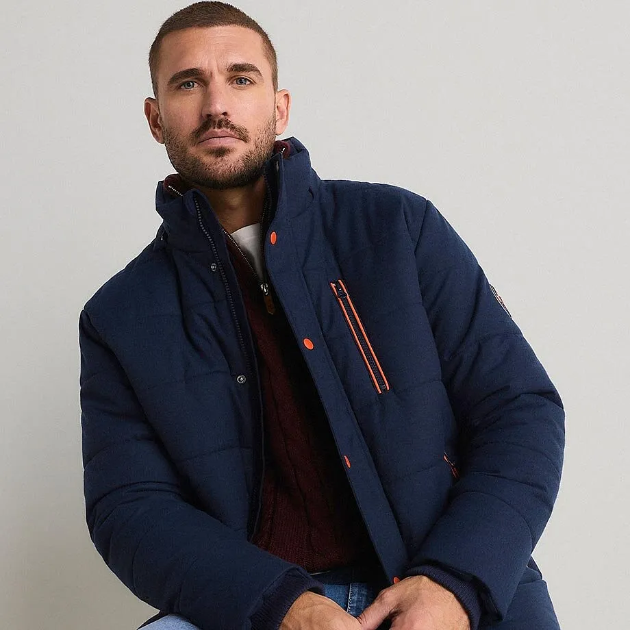 Mann tr&auml;gt eine dunkelblaue Steppjacke, einen weinroten Pullover und hellblaue Jeans.