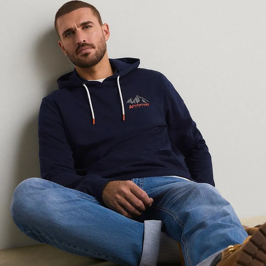 Er tr&auml;gt einen dunkelblauen Hoodie mit Bergmotiv und dazu eine l&auml;ssige blaue Jeans.