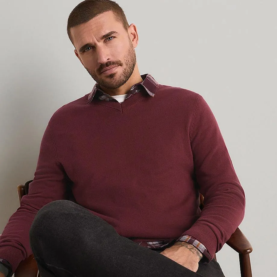 Mann tr&auml;gt einen bordeauxroten Pullover, ein kariertes Hemd und dunkle Jeans.