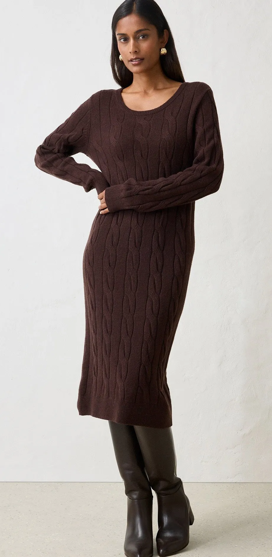 Elegantes braunes Strickkleid mit Zopfmuster, kombiniert mit hohen dunklen Stiefeln und Ohrringen.