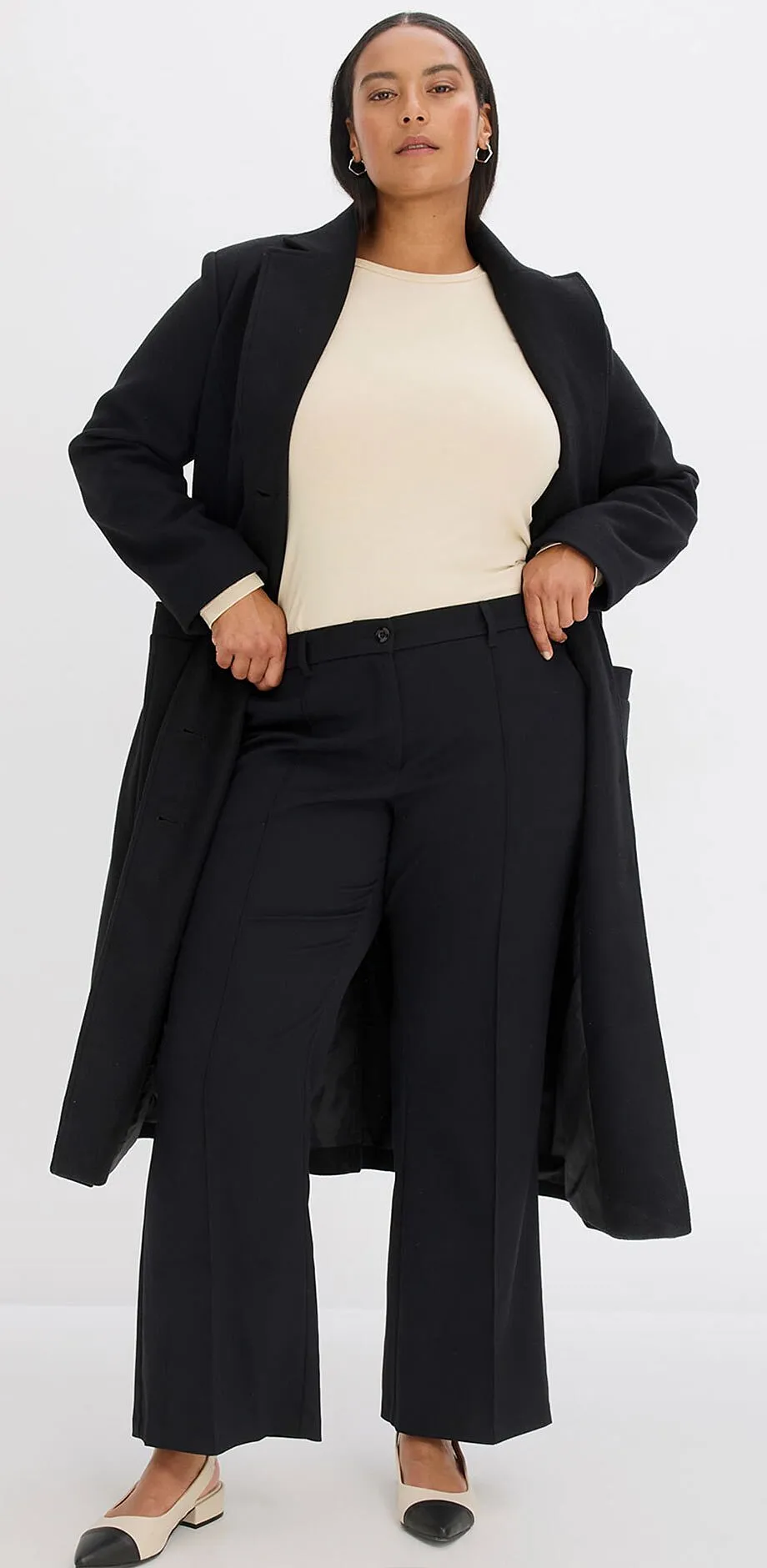 Manteau noir par dessus un pull couleur cr&egrave;me, associ&eacute; &agrave; un pantalon large noir.