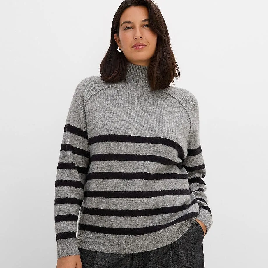 Gestrickter Rollkragenpullover in Grau mit schwarzen Querstreifen und dunkler Hose.